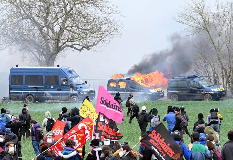A flaming police car in a field and protesters waving flags.