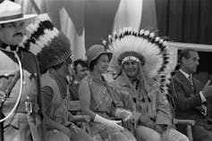 the Queen sits between two Indigenous leaders, each wearing a headress.