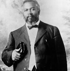 A man with facial hair poses, wearing a suit jacket, white shirt, and dark bow tie.
