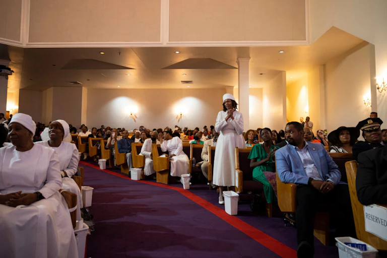 Rows of people, most of whom are Black and in formal clothing, sit in church pews inside a sanctuary with beige walls.