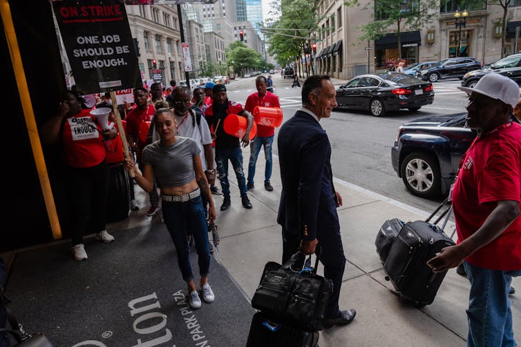 A group of strikers, including one holding a sign saying 'One job should be enough' sign aloft encounter a man in a suit and wheeled luggage.