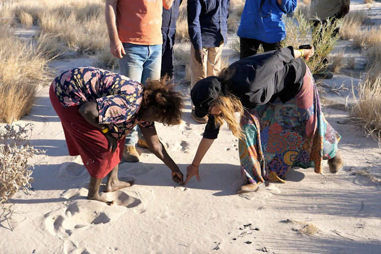 Two women point to marks in the desert sand made by animals