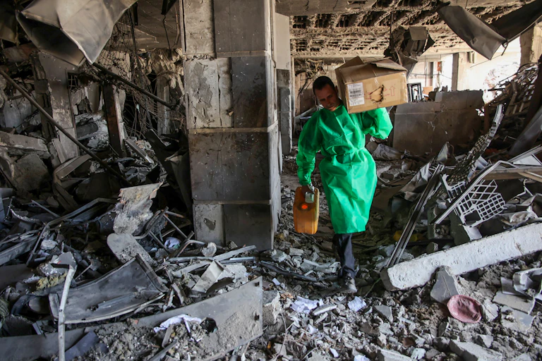 A man in a green smock carries a box through rubble.