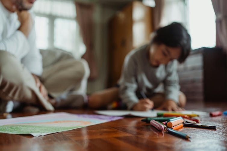 A young child and a man sit on the floor with colour pencils and papers. The child is drawing while the man looks on.