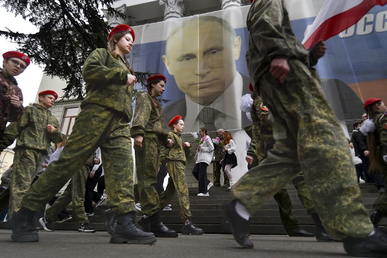 Young people wearing army fatigues and red hats march in a military parade.