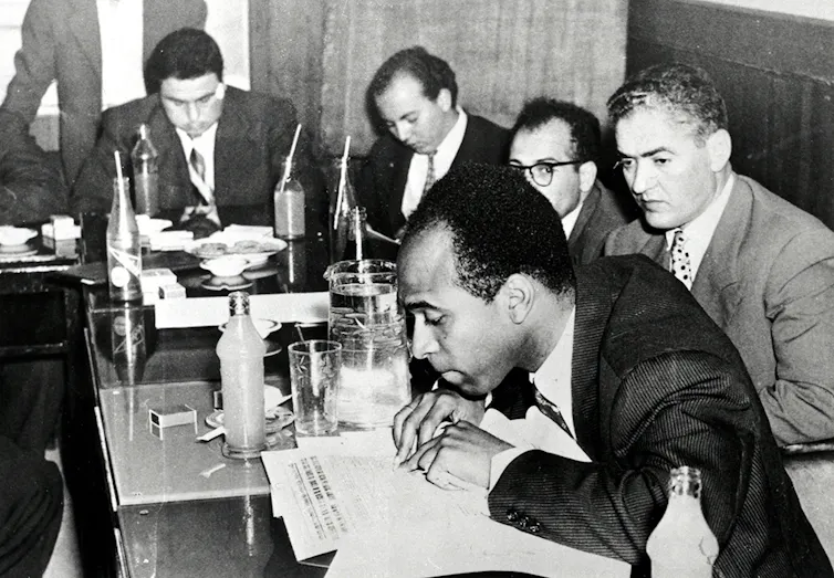 A black and white photograph of a group of men sitting at desks, in the foreground a Black man reads closely from a text.