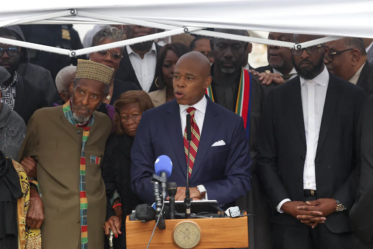 Adams speaks at a microphone surrounded by people