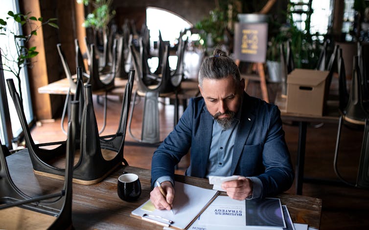 A man with gray hair in a bun, wearing a blue suit, sits doing paperwork at a table surrounded by other tables with the chairs placed on top of them.
