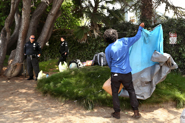 A man folds a tent as two uniformed police watch from a distance