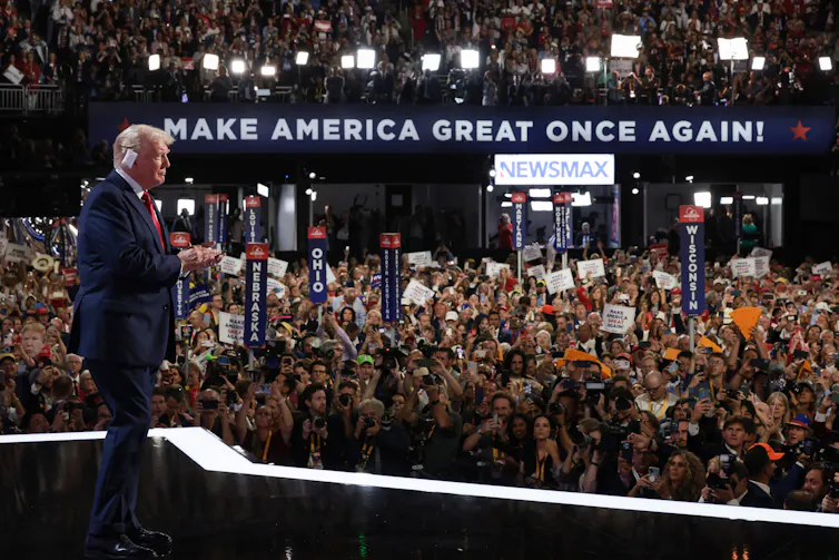 An older white man with a blue suit and red tie has a bandage over his ear and stands on a stage in front of a large crowd of people, with a banner that says 'Make America Great Again.'