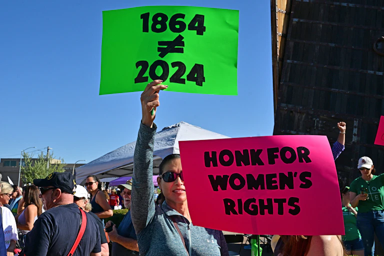 A woman holds up a neon pink sign that says 'Honk for women's rights' and a neon green one that says '1864 does not equal 2024'