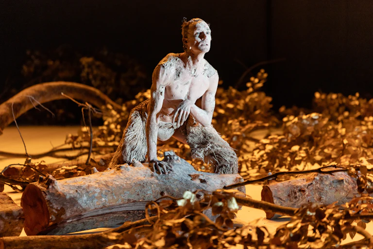 Production image: a man covered in mud leans on a tree stump.