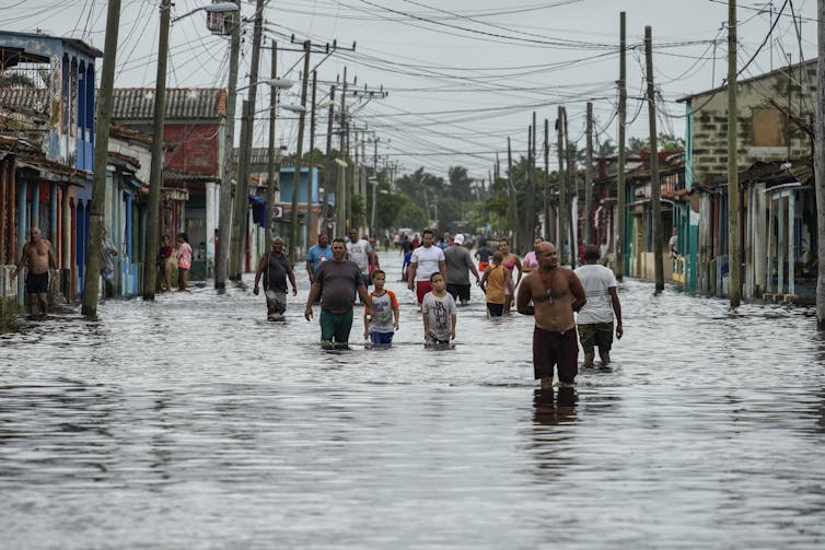 People walking through knee-deep water on a flooded street with building on either side and electric wires overhead.