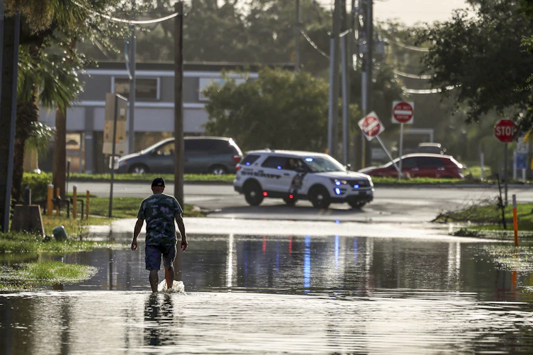 A man walks through ankle-deep water toward a police car at the end of the road. Road signs are knocked askew.