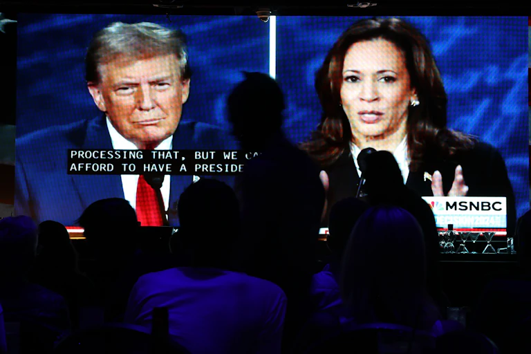 people in darkened room watching Trump and Harris on large screen during debate