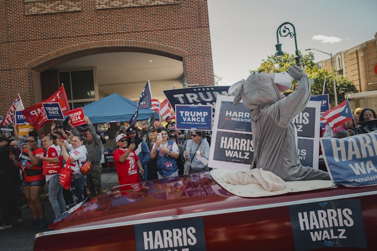 Red car with Harris-Walz signs drives past a crowd of people holding Trump-Vance signs and Puerto Rican flags