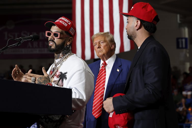 Man in red MAGA hat and funky sunglasses and wearing big chain around neck speaks at a podium with Donald Trump behind him