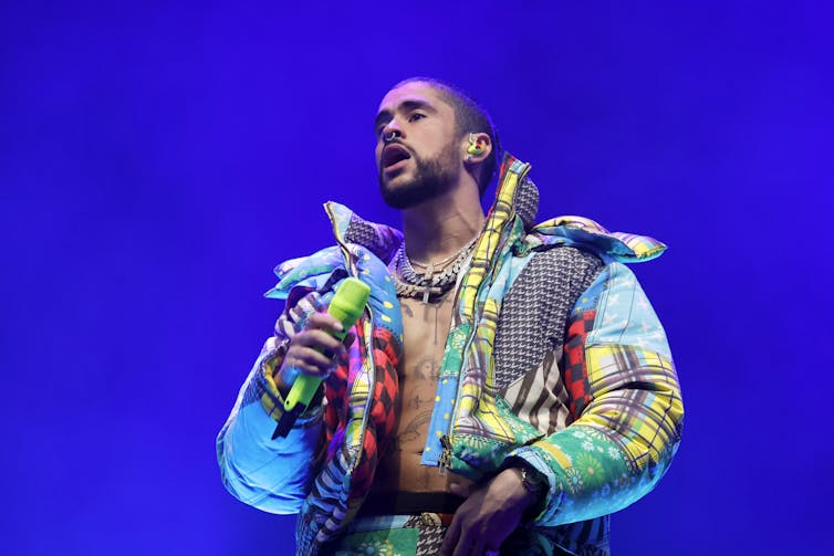 Man wearing multicolored and multipatterned puffer jacket and chunky necklaces holds neon green microphone