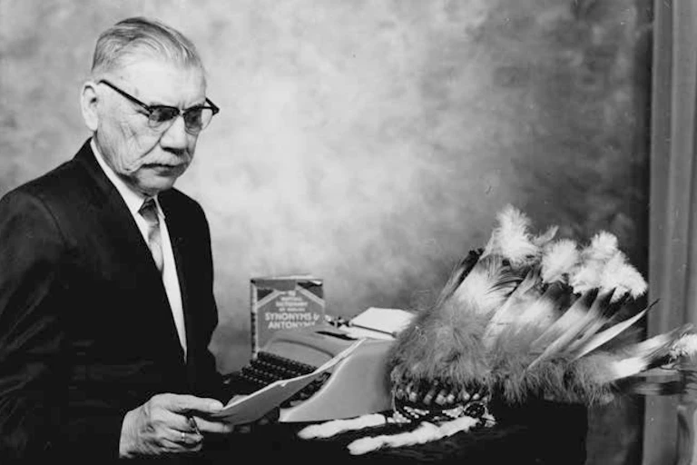 A man looking at papers, in a posed photo near a typewriter with a book and feathers.