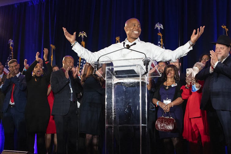 A jubiland man in a white shirt, arms raised in celebration, standing at a lectern with a crowd of happy people clapping behind him.