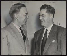 A black and white photo shows two men in suits smiling as they talk.