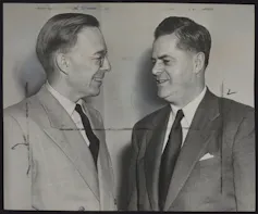 A black and white photo shows two men in suits smiling as they talk.