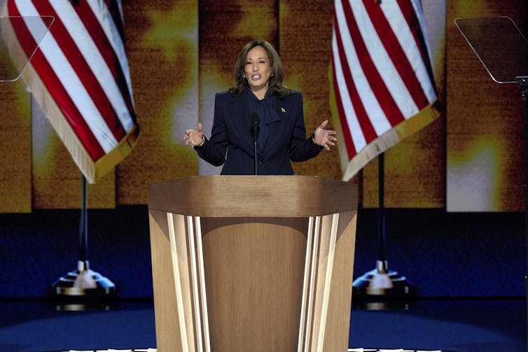 A woman in a suit speaks at a lectern, which has an American flag on either side.