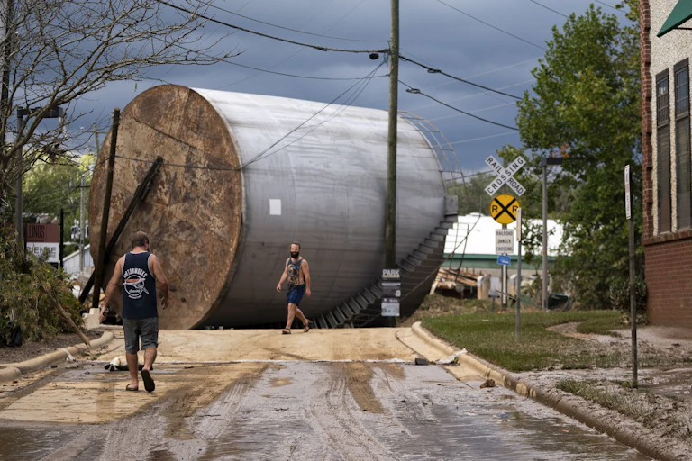 Two men walk near a giant storage tower that lies on its side near rail road tracks.