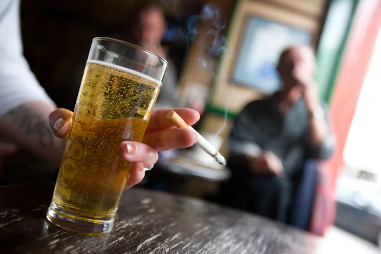 A person's hand holding a pint of beer and a lit cigarette.