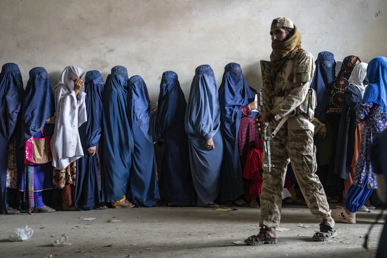 Um homem armado está ao lado de uma longa fila de mulheres cobertas da cabeça aos pés com um hijab.