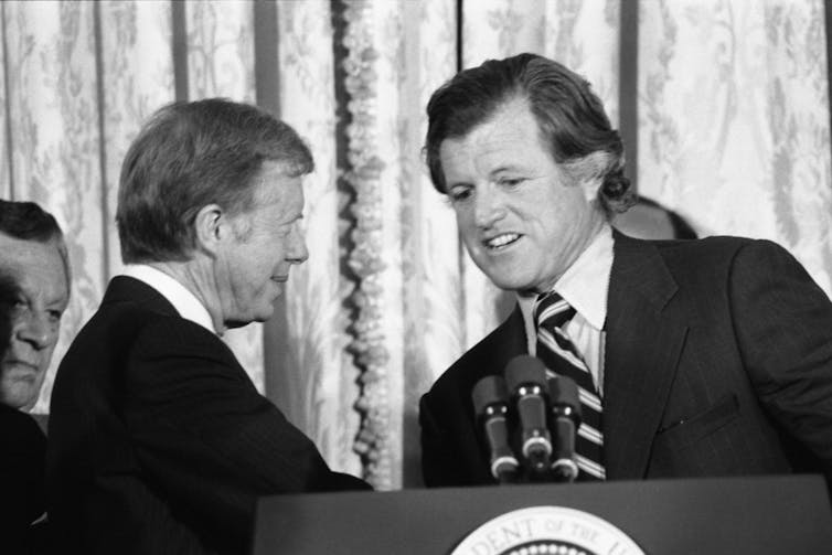 Two middle-aged white men dressed in business suits are shaking hands.