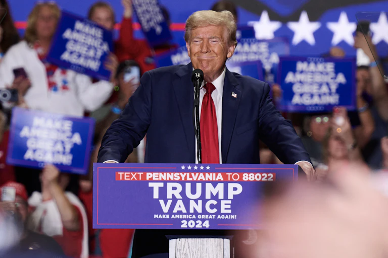 A man in a blue jacket, white shirt and red tie in front of a large crowd.