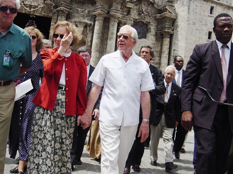 A middle aged white couple are walking and holding hands as they are surrounded by security officials.