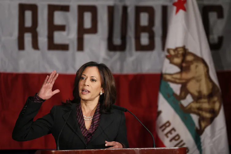 A woman gestures while standing in front of the California state flag.