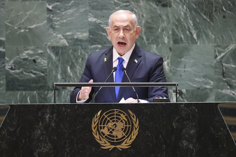 A man in a suit stands at a United Nations lectern and speaks