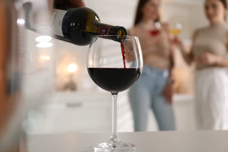 Photo de quelqu’un en train de servir un verre de vin en gros plan, avec à l’arrière-plan deux femmes.