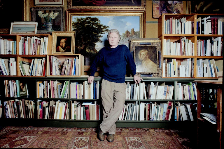 Brian Sewell in front of a large book case and a wall covered in paintings