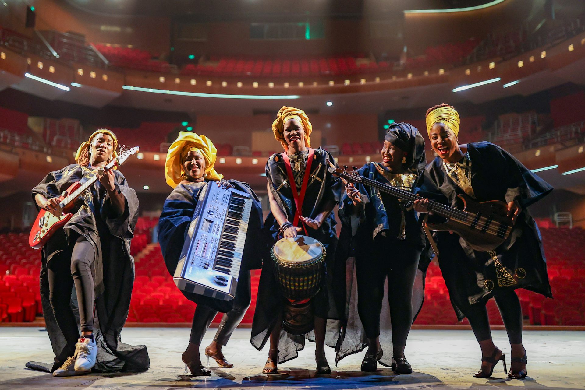 Five women on a stage, in a row, playing musical instruments and smiling, traditional cloth on their heads and flowing robes on.