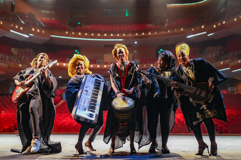 Five women on a stage, in a row, playing musical instruments and smiling, traditional cloth on their heads and flowing robes on.