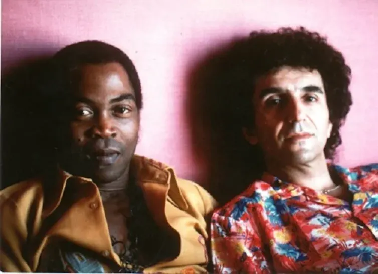 Two men lean against a red wall and look at the camera with half-smiles, dressed in vintage clothing.