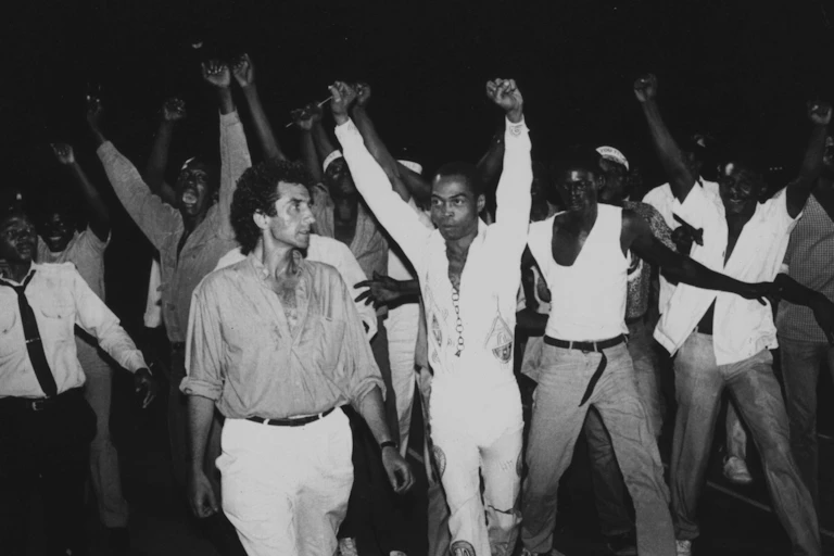 A black and white image of a crowd of men, jubilant and arms raised. A man with curly hair stands in front next to an African man with both arms raised in defiant fists.