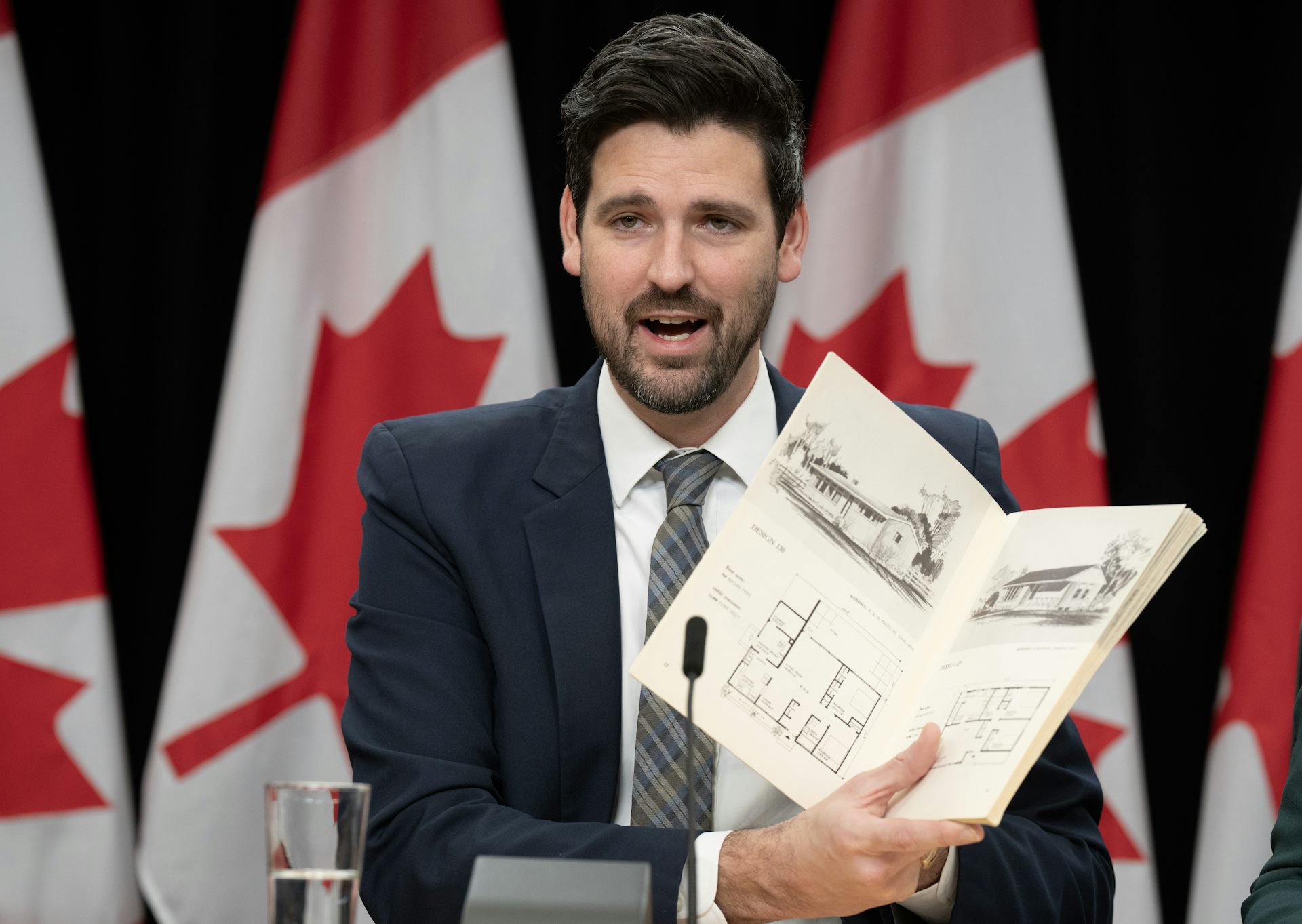 A middle-aged man holds up a booklet with sketches of houses and floor plans in it while seated in front of Canadian flags