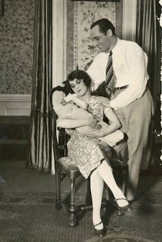 A black and white photograph of a man in office clothing smiling at a seated woman who holds an infant.