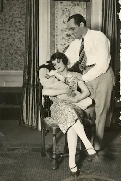 A black and white photograph of a man in office clothing smiling at a seated woman who holds an infant.
