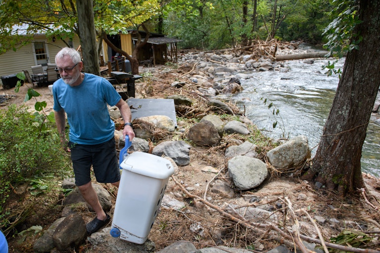 A man carries belonging out of a homes that flooded near a creek. Debris is washed up all around.
