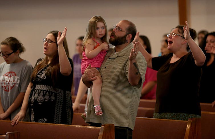 A man holds a toddler in a pink outfit as he and other people in church pews pray with their arms raised.