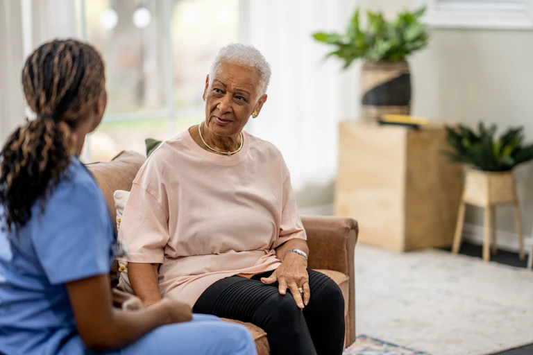 A young woman in scrubs visits with an older woman in a private home.