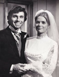 A black-and-white photo of a man in a black suit and striped tie, smiling and holding the hand of a blond woman in a white dress and veil.