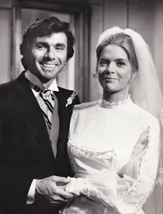 A black-and-white photo of a man in a black suit and striped tie, smiling and holding the hand of a blond woman in a white dress and veil.