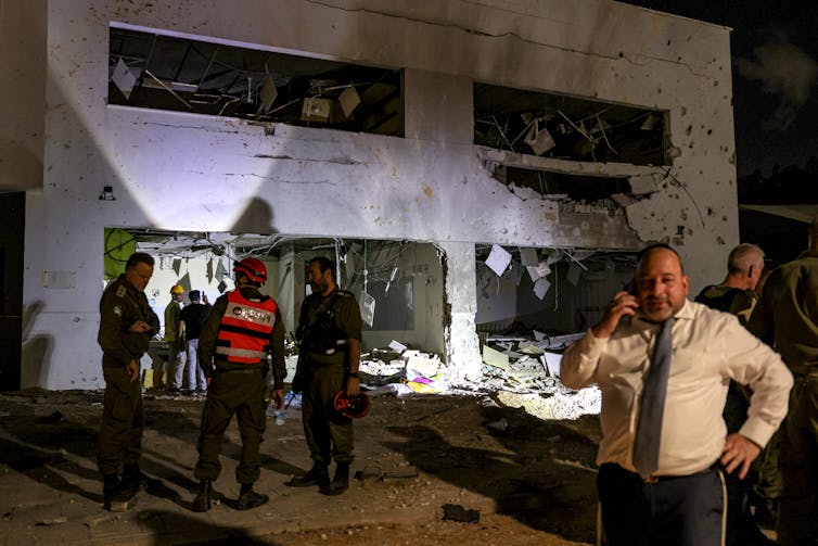 People stand around a gray building at nighttime that appears damaged.